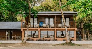 Two story wooden beach house with an upstairs balcony and tropical trees overhead outdoor seating on both levels