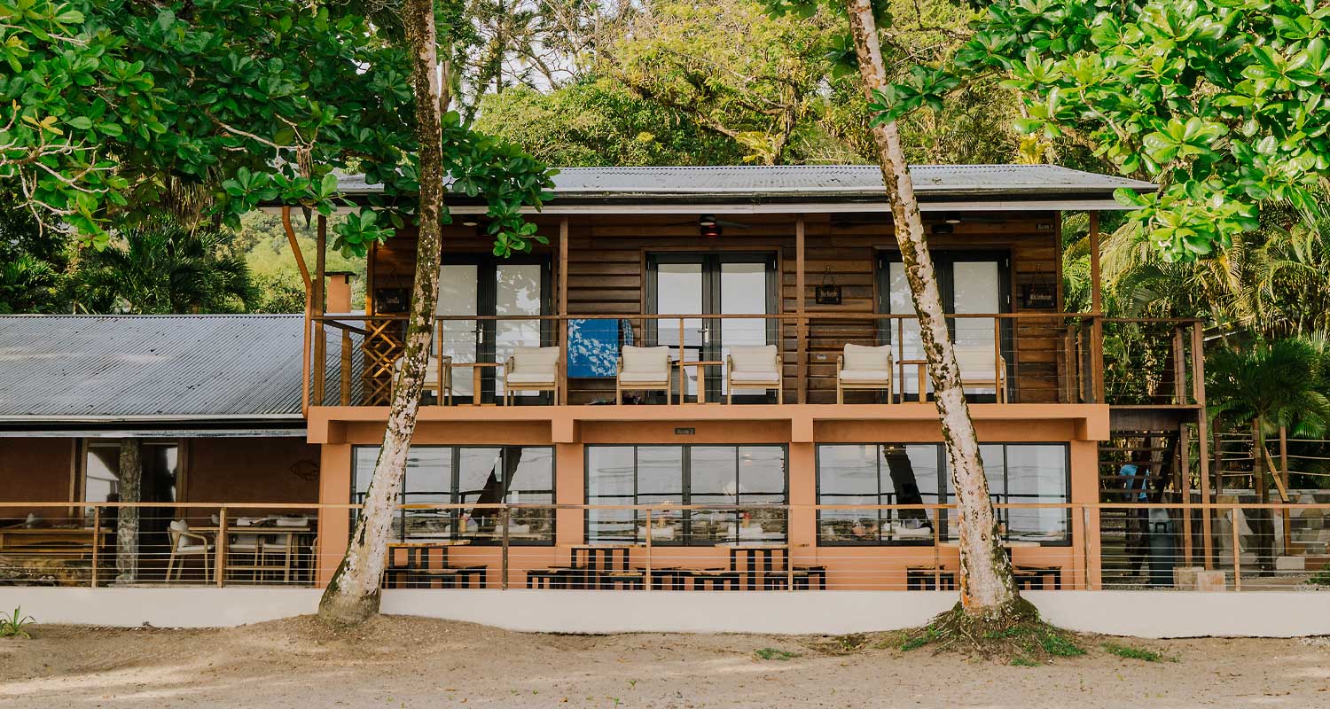 Two-story wooden beach house with an upstairs balcony and tropical trees overhead, outdoor seating on both levels.
