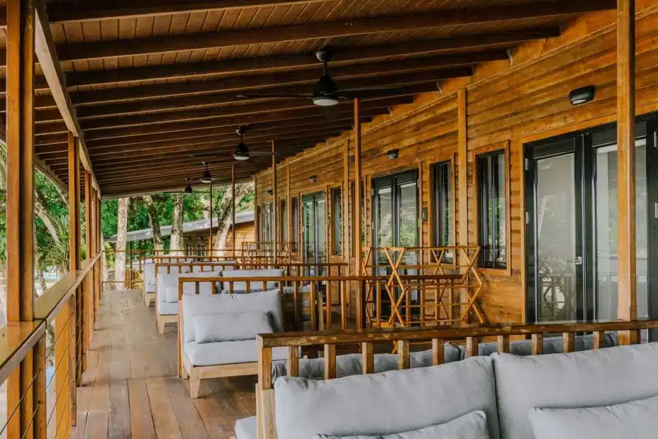 Wooden beachfront porch with row of cushioned benches facing a cabin, overlooking palm trees and sand beyond the railing.