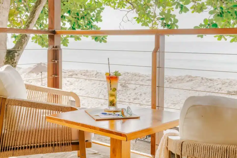 Seaside balcony table with a tall cocktail, sunglasses, notebook, and wicker chairs overlooking the beach and ocean.