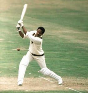 Cricketer in white batting mid swing on a cricket pitch with a ball approaching the bat